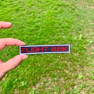 May include: A black and white patch with the text "FLIGHT RISK" in red embroidery.