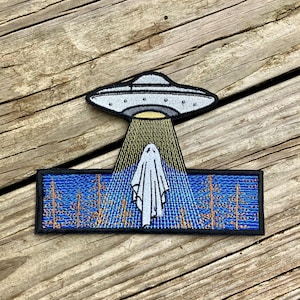 May include: Embroidered patch depicting a white ghost being abducted by a grey UFO. The ghost is standing in a blue and brown forest scene.