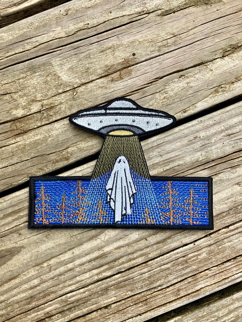 May include: Embroidered patch featuring a white ghost being abducted by a grey UFO. The ghost is standing in a blue and brown forest scene.
