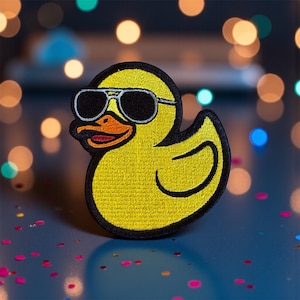 May include: Embroidered yellow duck patch wearing sunglasses. The duck has a black outline, orange beak, and a red tongue. The sunglasses are silver-framed with black lenses. The patch is on a blue surface with colorful confetti.
