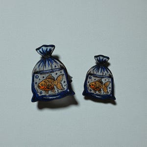 Puede incluir: Dos parches bordados con forma de bolsas azules, cada una con un pez dorado. Los peces son de color naranja con escamas detalladas. Las bolsas tienen un contorno azul oscuro y un fondo blanco punteado, lo que sugiere agua. Para ropa o accesorios.