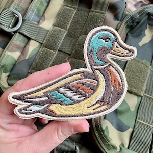 Może przedstawiać: Haftowana naszywka z kaczką z precyzyjnym wzorem w kolorach turkusowym, brązowym i żółtym. Naszywka w kształcie kaczki ma kremową obwódkę. Tło to wzór kamuflażu.