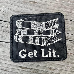 May include: Black square patch with a white embroidered design of three stacked books. The phrase "Get Lit." is embroidered in white below the books. The patch has a black border.