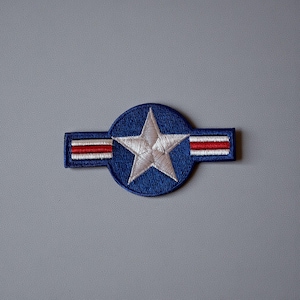 Può includere: Toppa ricamata con una stella bianca all'interno di un cerchio blu, affiancata da rettangoli blu con strisce rosse e bianche. La toppa ha un aspetto strutturato ed è probabilmente progettata per essere applicata su abbigliamento o accessori.