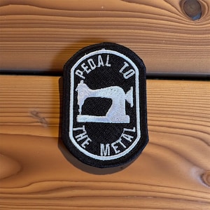 May include: Black and white embroidered patch with the text "PEDAL TO THE METAL" surrounding a white sewing machine silhouette. The patch has a black border and is set against a wooden background.