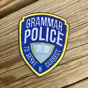 Puede incluir: Un parche bordado azul y amarillo con el texto "Grammar Police" y "To Serve & Correct". El parche presenta una imagen estilizada de un libro abierto.
