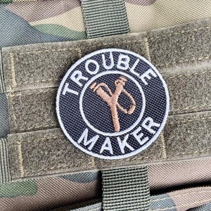 May include: Circular patch with the text "TROUBLE MAKER" in white around a navy blue circle. A brown slingshot graphic is in the centre. The patch is attached to camouflage fabric.