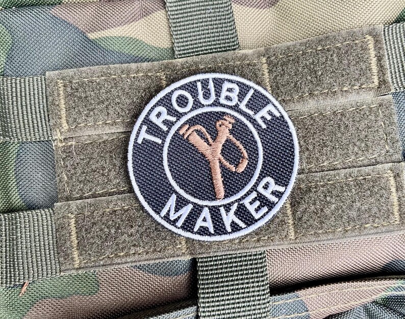 May include: Circular patch with the text "TROUBLE MAKER" in white around a navy blue circle. A brown slingshot graphic is in the centre. The patch is attached to camouflage fabric.