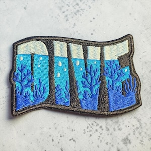 Peut inclure: Écusson brodé avec le mot "DIVE" en blanc, avec une scène sous-marine bleue à l'intérieur de chaque lettre. Les lettres sont soulignées en noir, avec une bordure marron. La scène sous-marine comprend du corail bleu et des bulles.