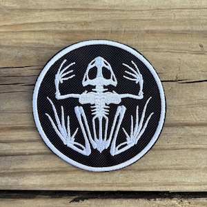 May include: A round, black patch with a white frog skeleton design. The skeleton is centered within the patch, with the bones detailed against the black background. The patch has a white border.