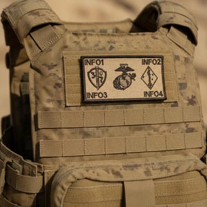May include: A tan camouflage tactical vest featuring a rectangular morale patch with black embroidered details. The patch displays the text INFO1, INFO2, INFO3, and INFO4, alongside a central Marine Corps emblem, a shield with a sword, and a diamond.
