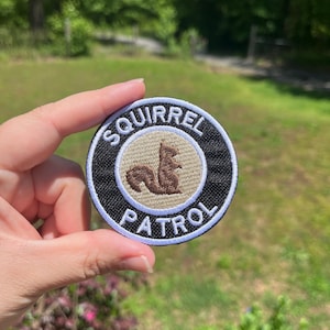Könnte beinhalten: Runder Aufnäher mit dem Text "SQUIRREL PATROL" in Weiß auf schwarzem Hintergrund. Ein braunes Eichhörnchen befindet sich in der Mitte eines beigen Kreises. Der Aufnäher wird vor einem verschwommenen grünen Hintergrund gehalten.