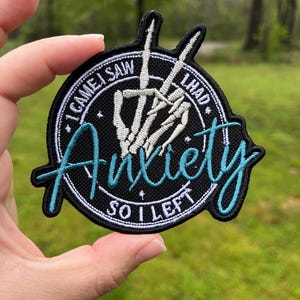 Pode incluir: Um remendo bordado preto e branco com uma mão de esqueleto fazendo o sinal de paz. O texto no remendo diz "Eu vim, eu vi, eu tive ansiedade, então eu fui embora".