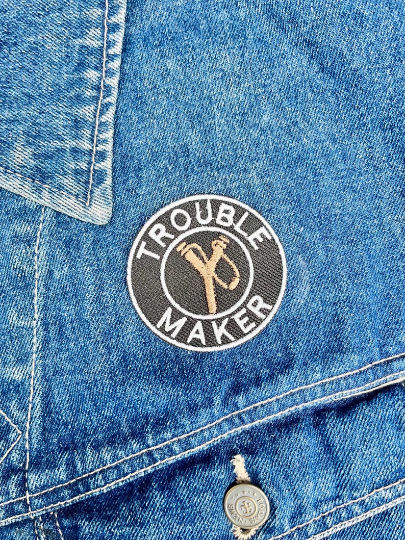 May include: Close-up of a blue denim jacket with white stitching. A circular patch with the words "TROUBLE MAKER" in white surrounds a gold and black graphic. A silver button is visible.