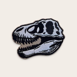 Puede incluir: Un parche bordado con un diseño detallado de un cráneo de dinosaurio en negro y gris claro. El parche muestra la estructura esquelética de una cabeza de T-Rex de perfil, con dientes y cuencas oculares prominentes en hilos de colores.