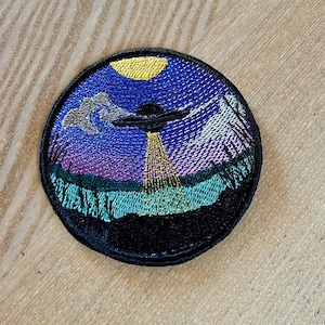 Puede incluir: Parche bordado con una escena de ovni. Un platillo volante negro se cierne sobre un paisaje con un cielo azul y púrpura, una luna amarilla y follaje verde y negro. El parche tiene un borde negro.