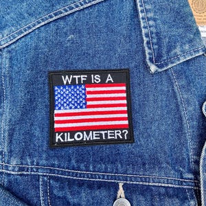 Pode incluir: Uma jaqueta de ganga azul com um remendo preto que diz "WTF IS A KILOMETER?" e apresenta uma bandeira americana.