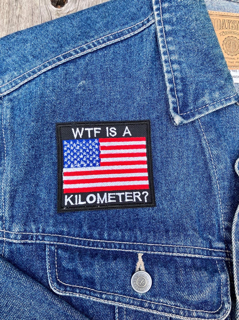 Pode incluir: Uma jaqueta de ganga azul com um remendo preto que diz "WTF IS A KILOMETER?" e apresenta uma bandeira americana.