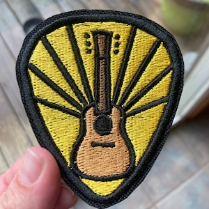 May include: A yellow and brown embroidered patch of a guitar with sun rays behind it. The patch is surrounded by a black border.
