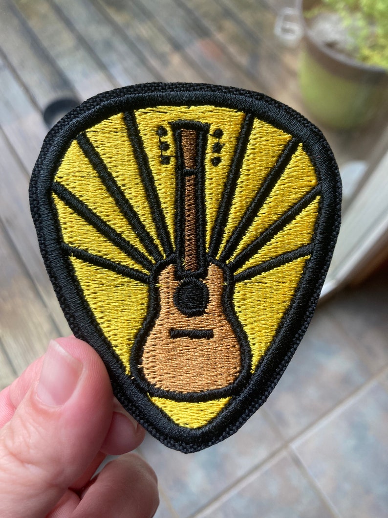 Może przedstawiać: Naszywkę haftowaną w kolorze ż&oacute;łtym i brązowym przedstawiającą gitarę z promieniami słońca za nią. Naszywka jest otoczona czarną obw&oacute;dką.