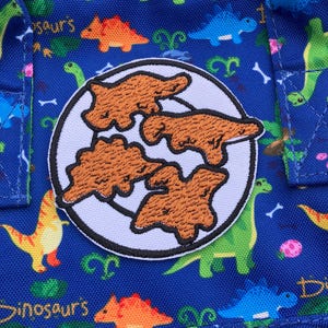 May include: A blue fabric bag with a white circle patch featuring three brown embroidered dinosaurs. The bag is covered in a repeating pattern of colorful dinosaurs.
