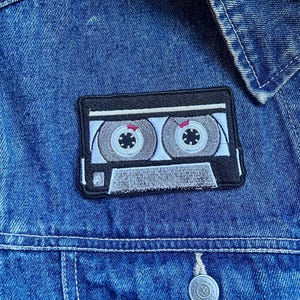 Pode incluir: Uma jaqueta de ganga azul com um remendo bordado em preto e branco que representa uma fita cassete. O remendo tem um fundo branco com detalhes pretos e duas bobinas prateadas.