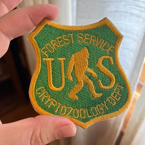 May include: An embroidered patch in green and gold with the text "US Forest Service Cryptozoology Dept." and a silhouette of Bigfoot.