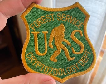 Puede incluir: Un parche bordado verde y dorado con el texto "US Forest Service Cryptozoology Dept." y una silueta de Bigfoot.