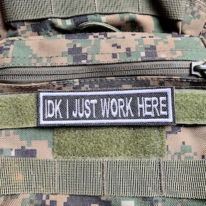 May include: A black and white patch with white stitching that reads "IDK I JUST WORK HERE" is attached to a green and brown camouflage fabric.