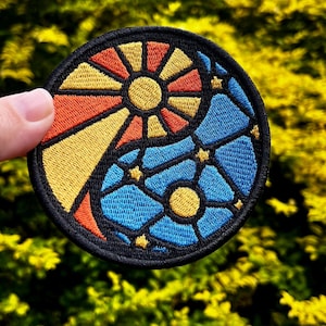 Könnte beinhalten: Runder, gestickter Aufnäher mit einem Buntglas-Design. Er ist in zwei Bereiche unterteilt: einen gelb-orangenen Sonnenaufgang und einen blauen Bereich mit einem gelben Mond und Sternen. Der Aufnäher hat einen schwarzen Rand und einen grünen Hintergrund.