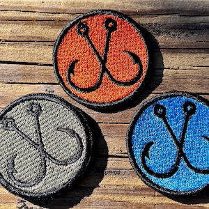 May include: Three round embroidered patches with a fishing hook design. One patch is orange, one is blue, and one is gray. Each patch has a black border and a black hook design. The patches are on a wooden surface.