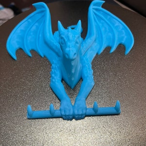 Pode incluir: Um gancho de parede de dragão azul claro com três ganchos. O dragão está em posição de agachamento com as asas abertas.