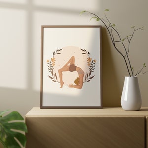 May include: A framed art print featuring a yoga pose illustration. The artwork depicts a person in a bridge pose, surrounded by a floral wreath. The color palette includes warm tones of brown, beige, and orange. The print is displayed on a wooden cabinet.