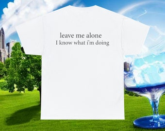 Può includere: T-shirt bianca con la scritta "leave me alone I know what I'm doing" in nero, con un font semplice. La maglietta è presentata su uno sfondo di erba verde, cielo blu e skyline.