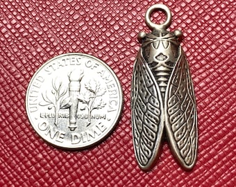 May include: A silver-toned cicada pendant with detailed wing patterns, next to a United States one dime coin. The pendant has a loop for hanging. The background is a textured, red surface.