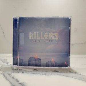 May include: A CD case with a blue background and a white, pixelated band name "The Killers" and the album title "Hot Fuss" in red text.