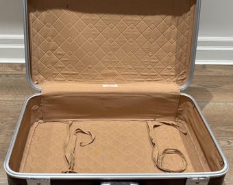 Puede incluir: Maleta vintage abierta, marrón con ribete plateado. El interior presenta un forro acolchado beige y una solapa beige con una pequeña etiqueta. Incluye correas beige. El asa es marrón.