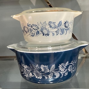 Puede incluir: Dos fuentes de horno Pyrex vintage con tapas. La fuente superior es blanca con diseños florales azules, y la fuente inferior es azul marino con diseños florales blancos. Ambas fuentes tienen tapas de vidrio transparente y asas laterales.