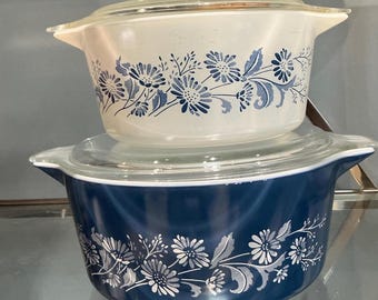 May include: Two vintage Pyrex casserole dishes with lids. The top dish is white with blue floral designs, and the bottom dish is navy blue with white floral designs. Both dishes have clear glass lids and side handles.
