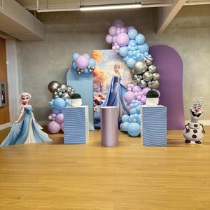 May include: A themed party setup with Frozen decorations. The scene includes a backdrop with Elsa, balloon arches in blue, silver, and purple, and decorative pedestals. A cutout of Olaf the snowman is also present.
