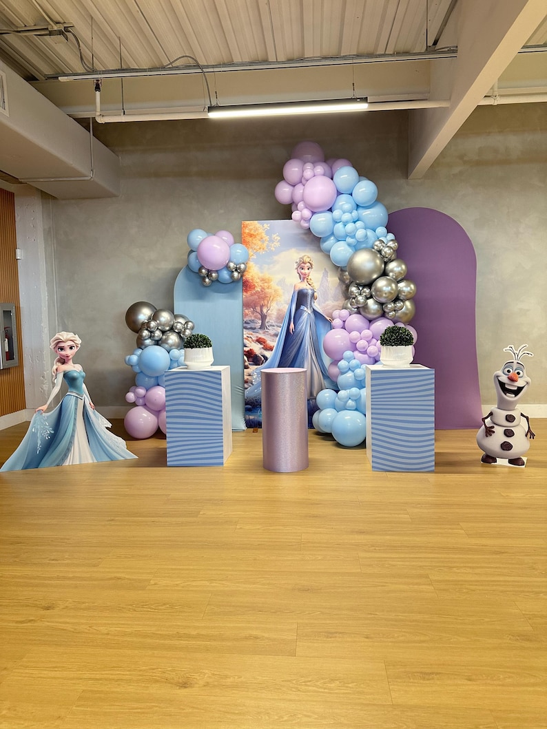 May include: A themed party setup with Frozen decorations. The scene includes a backdrop with Elsa, balloon arches in blue, silver, and purple, and decorative pedestals. A cutout of Olaf the snowman is also present.