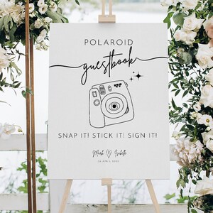 以下が含まれることがあります： 「POLAROID guestbook」と黒文字で書かれた白い看板。ポラロイドカメラの絵と「SNAP IT! STICK IT! SIGN IT!」の文字が書かれている。看板には「Mark & Grabelle」の名前と「04 APRIL 2023」の日付も書かれている。