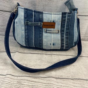 May include: A shoulder bag constructed from vertical strips of light and dark blue denim fabric. The bag features a dark blue denim strap, silver-toned metal hardware, and a brown rectangular leather label with the text Handmade By Lucille B.