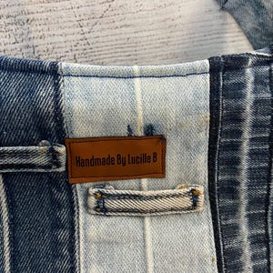 May include: A patchwork denim garment featuring alternating panels of dark and light blue wash fabric. A rectangular brown leather label is stitched onto the center panel, displaying the black text "Handmade By Lucille B" with a belt loop below.