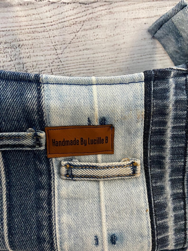 May include: A patchwork denim garment featuring alternating panels of dark and light blue wash fabric. A rectangular brown leather label is stitched onto the center panel, displaying the black text "Handmade By Lucille B" with a belt loop below.