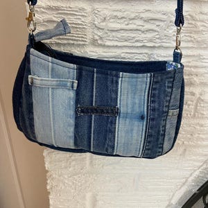 May include: A small crossbody bag constructed from vertical strips of denim fabric in varying shades of blue, ranging from light wash to dark indigo. The bag features a zippered top closure, silver-toned metal hardware, and a matching denim strap.
