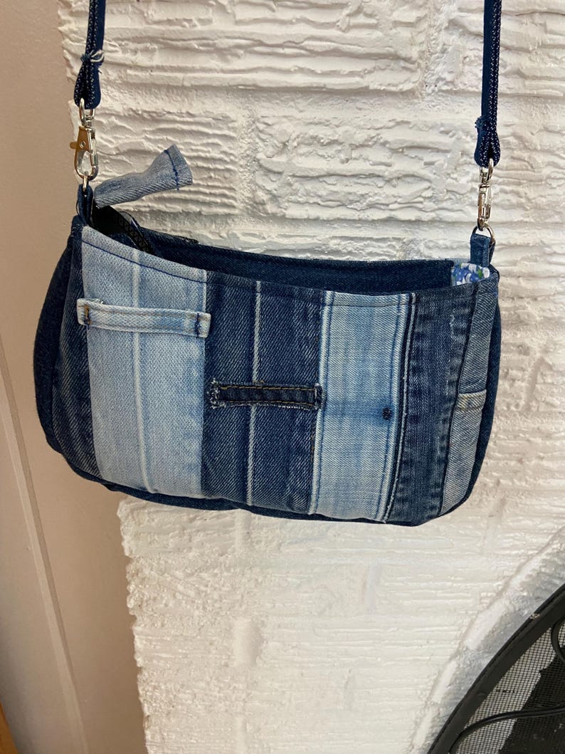 May include: A small crossbody bag constructed from vertical strips of denim fabric in varying shades of blue, ranging from light wash to dark indigo. The bag features a zippered top closure, silver-toned metal hardware, and a matching denim strap.