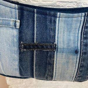 May include: A patchwork bag constructed from various shades of blue denim fabric. The design features vertical panels of light and dark wash denim, including a visible horizontal belt loop detail stitched onto the center of the fabric surface.