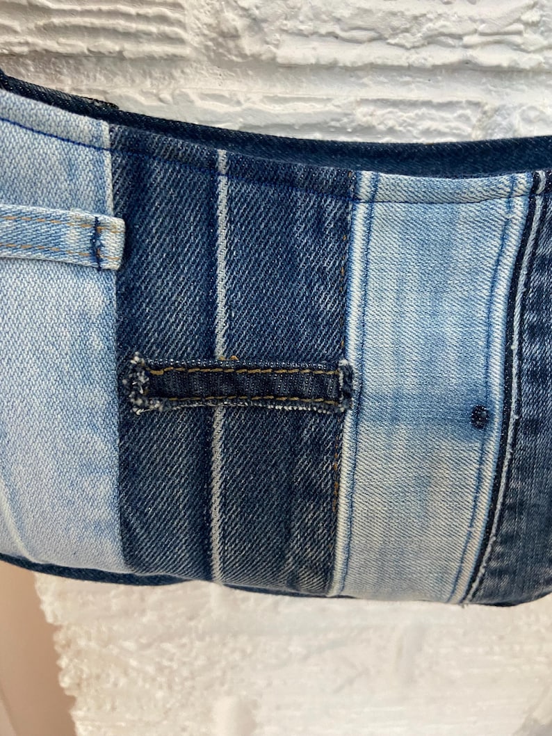 May include: A patchwork bag constructed from various shades of blue denim fabric. The design features vertical panels of light and dark wash denim, including a visible horizontal belt loop detail stitched onto the center of the fabric surface.