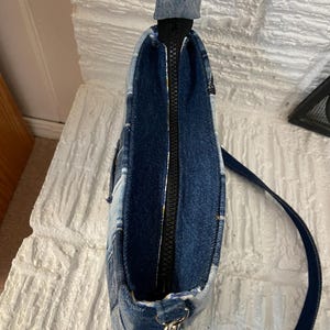 May include: A shoulder bag constructed from various shades of blue denim fabric patches. The bag features a top zipper closure with a black zipper track, a single denim strap attached with silver-toned metal hardware, and a visible interior lining.